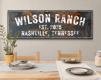 May include: A rectangular wooden sign with the words "WILSON RANCH EST. 2025 NASHVILLE, TENNESSEE" in white lettering. The sign has a distressed, textured background with dark and gold tones. The sign is displayed above a dining table.