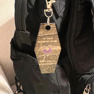 Op de afbeelding: Houten sleutelhanger met een goudkleurige sluiting. De sleutelhanger heeft een geometrische vorm en bevat de tekst "IN THIS MOMENT NOW, CAPTURE IT, REMEMBER IT." Een paars hart staat in het midden van het ontwerp.