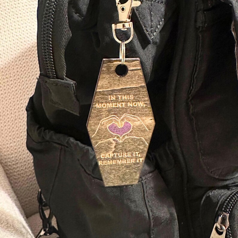 Op de afbeelding: Houten sleutelhanger met een goudkleurige sluiting. De sleutelhanger heeft een geometrische vorm en bevat de tekst "IN THIS MOMENT NOW, CAPTURE IT, REMEMBER IT." Een paars hart staat in het midden van het ontwerp.