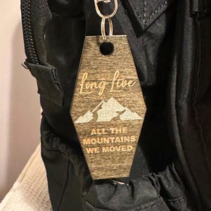 Op de afbeelding: Een houten sleutelhanger met een bergontwerp en de woorden "Long Live All The Mountains We Moved" in een lichte kleur gegraveerd. De sleutelhanger is bevestigd aan een zilveren sleutelring en hangt aan een zwarte rugzak.