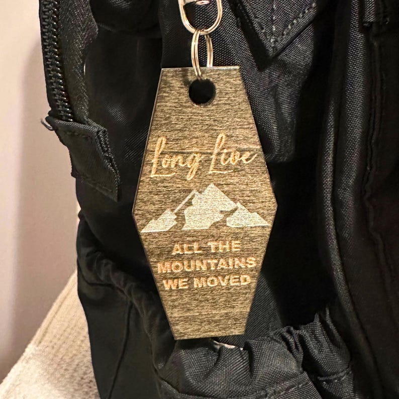 Op de afbeelding: Een houten sleutelhanger met een bergontwerp en de woorden "Long Live All The Mountains We Moved" in een lichte kleur gegraveerd. De sleutelhanger is bevestigd aan een zilveren sleutelring en hangt aan een zwarte rugzak.