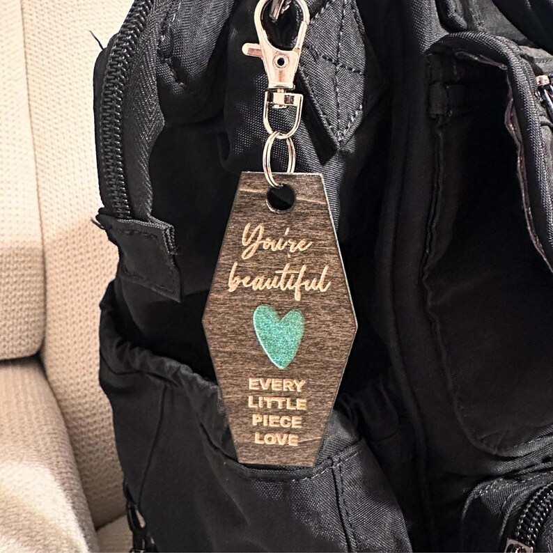 Op de afbeelding: Houten sleutelhanger met de woorden "You're beautiful", een turquoise hart en "Every little piece love" gegraveerd. De sleutelhanger is bevestigd aan een zilveren clip. De sleutelhanger hangt aan een zwarte tas.