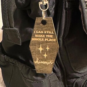 Op de afbeelding: Een houten sleutelhanger met een zilverkleurige sluiting. De sleutelhanger is een lichtbruin, doodskistvormig label met de woorden "I CAN STILL MAKE THE WHOLE PLACE" en het woord "shimmer" gegraveerd, samen met drie sterrenburst-ontwerpen.