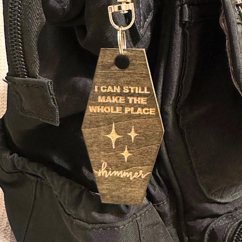 Op de afbeelding: Een houten sleutelhanger met een zilverkleurige sluiting. De sleutelhanger is een lichtbruin, doodskistvormig label met de woorden "I CAN STILL MAKE THE WHOLE PLACE" en het woord "shimmer" gegraveerd, samen met drie sterrenburst-ontwerpen.