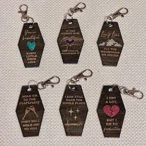 Op de afbeelding: Zes houten sleutelhangers met zilveren sluitingen. Elke sleutelhanger heeft een uniek ontwerp en tekst, waaronder zinnen als "You're beautiful", "Long live" en "Hold on to the memories". De sleutelhangers hebben verschillende illustraties en kleuren.