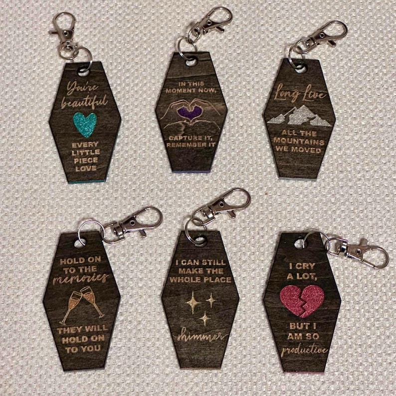Op de afbeelding: Zes houten sleutelhangers met zilveren sluitingen. Elke sleutelhanger heeft een uniek ontwerp en tekst, waaronder zinnen als "You're beautiful", "Long live" en "Hold on to the memories". De sleutelhangers hebben verschillende illustraties en kleuren.