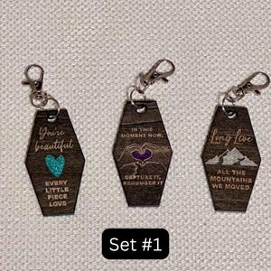 Op de afbeelding: Set van drie houten sleutelhangers met zilveren sluitingen. Elke sleutelhanger heeft een uniek ontwerp en tekst: "You're beautiful", een turquoise hart en "Every little piece love"; "In this moment now, capture it, remember it"; en "Long live, all the mountains we moved."