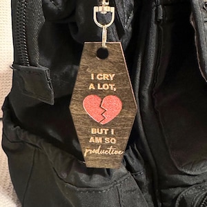 Op de afbeelding: Houten sleutelhanger met een zilverkleurige metalen sluiting. De sleutelhanger is in de vorm van een doodskist en heeft de tekst "I CRY A LOT, BUT I AM SO productive" met een rode gebroken hartafbeelding. De sleutelhanger is bevestigd aan een zwarte rugzak.