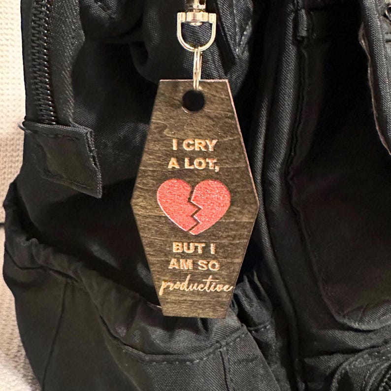 Op de afbeelding: Houten sleutelhanger met een zilverkleurige metalen sluiting. De sleutelhanger is in de vorm van een doodskist en heeft de tekst "I CRY A LOT, BUT I AM SO productive" met een rode gebroken hartafbeelding. De sleutelhanger is bevestigd aan een zwarte rugzak.