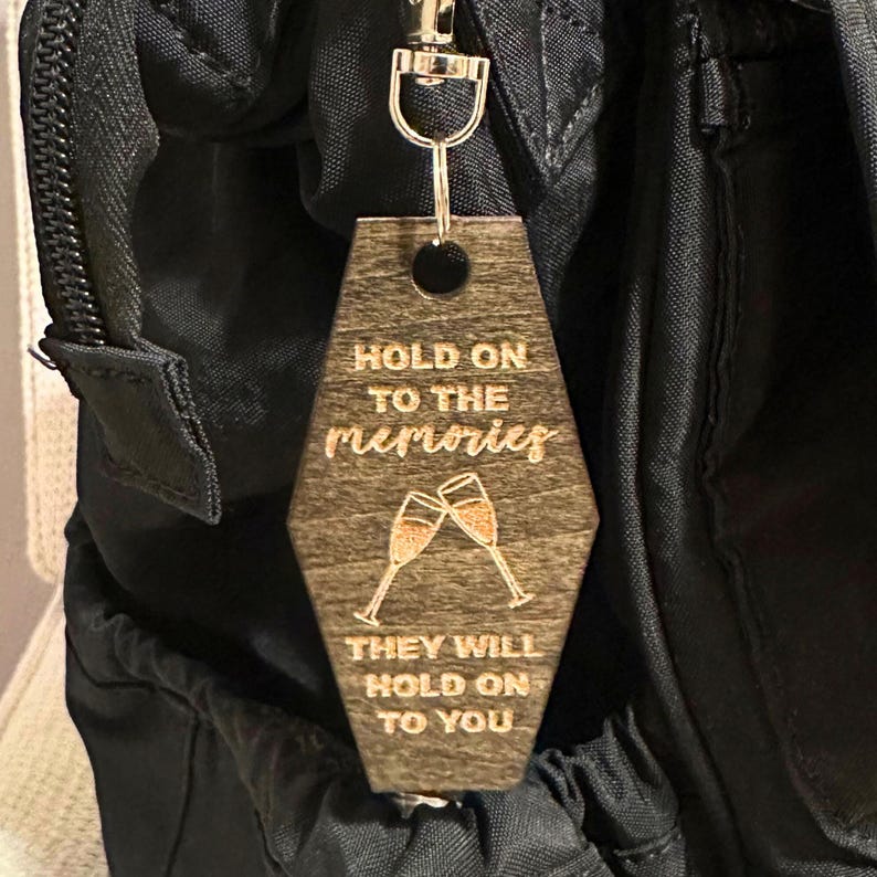 Op de afbeelding: Houten sleutelhanger met een donkerbruine afwerking, met de gegraveerde tekst "HOLD ON TO THE memories THEY WILL HOLD ON TO YOU" en twee champagneglazen. De sleutelhanger is bevestigd aan een zilveren rits.