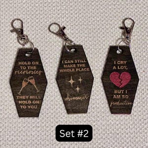 Op de afbeelding: Set van drie houten sleutelhangers met zilverkleurige sluitingen. Elke sleutelhanger, in de vorm van een kist, is gegraveerd met tekst en afbeeldingen, waaronder champagneglazen, sterren en een gebroken hart. De tekst bevat "Hold on to the memories", "I can still make the whole place shimmer" en "I cry a lot, but I am so productive."