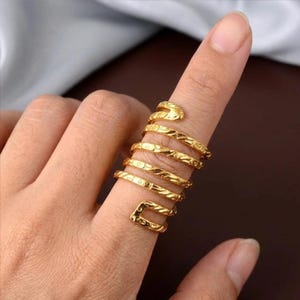 Może przedstawiać: Złoty pierścionek w kształcie węża owinięty wokół palca. Pierścionek ma teksturowany wzór i głowę węża na górze. Pierścionek ma spiralny kształt i jest wykonany z błyszczącego metalu.