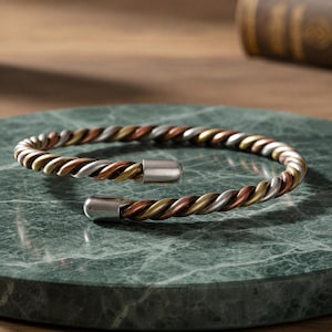 Puede incluir: Una pulsera de brazalete de metal trenzado tricolor con hebras alternas en tonos cobre, oro y plata. El diseño abierto está rematado con tapas cilíndricas lisas y pulidas de tono plateado en cada extremo de la banda de metal trenzado.