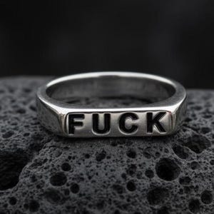 May include: A silver-colored ring with the word "FUCK" embossed in black capital letters. The ring has a flat, rectangular design and is set against a dark, textured background.