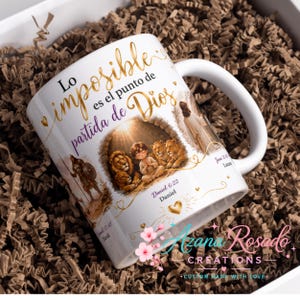 Puede incluir: Una taza de cerámica blanca con una ilustración religiosa de una persona sentada con leones dentro de una estructura de piedra. El texto dice Lo imposible es el punto de partida de Dios, Daniel 6:22 y Samuel 17:47. También se ven corazones dorados y una figura con túnica.