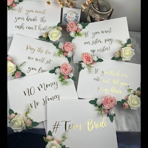 May include: Five white signs with gold lettering and pink and white flowers attached. The signs read: "If you want your bride the cash can't hide", "Pay the fee we will set you free", "No Money No Honey", "If you want our sister, pay up mister!" and "If you think I'm cute wait till you see my khala". The signs are arranged in a semi-circle with the sign that reads "#Team Bride" in the centre.