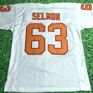 May include: White football jersey with orange and red trim. The number "63" and the name "SELMON" are printed on the back of the jersey.