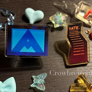 Może przedstawiać: Kolekcja małych, kolorowych akrylowych zawieszek i ozdób. Zawiera niebieskie serce, gwiazdę, kokardę i prostokątną zawieszkę z niebieskim geometrycznym wzorem. Jedna zawieszka głosi "HATE."