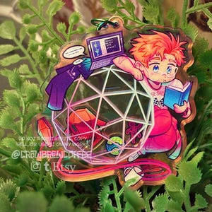 May include: A clear acrylic charm featuring an illustrated character with orange hair reading a blue book next to a laptop displaying the text REACH FOR THE STARS. A large geometric icosahedron shape contains smaller icons, including a red crystal and a globe.