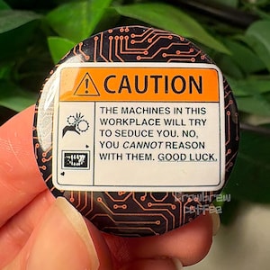 May include: A round pinback button featuring a black background with an orange circuit board pattern. The center displays a rectangular caution sign with the text: CAUTION THE MACHINES IN THIS WORKPLACE WILL TRY TO SEDUCE YOU. NO, YOU CANNOT REASON WITH THEM. GOOD LUCK.