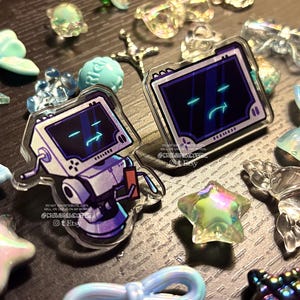 May include: Two acrylic pins featuring a cartoon robot with a screen face displaying a blue glowing expression. One pin shows the full robot body, while the other shows only the screen. They are surrounded by various small, translucent, light blue charms.