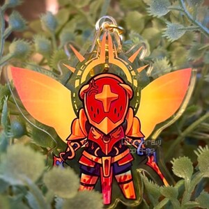 May include: A clear acrylic keychain featuring a stylized knight in red and gold armor with a cross emblem. The knight has orange wings and is surrounded by a gold halo. The keychain is hanging from a gold chain, set against a backdrop of green foliage.
