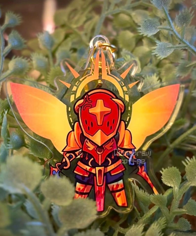 May include: A clear acrylic keychain featuring a stylized knight in red and gold armor with a cross emblem. The knight has orange wings and is surrounded by a gold halo. The keychain is hanging from a gold chain, set against a backdrop of green foliage.