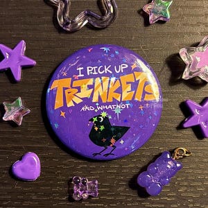 May include: A purple button with the text "I PICK UP TRINKETS AND WHATNOT" and a black bird illustration. The image also includes purple star and heart-shaped embellishments, and a purple gummy bear charm.