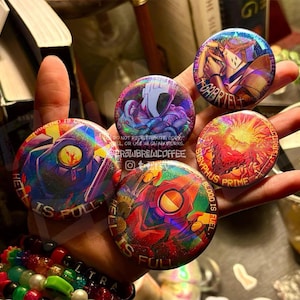 May include: Five circular pin-back buttons featuring holographic, colourful illustrations of characters and text including "Hell is full," "Gabriel," and "Sisyphus Prime." The designs incorporate vibrant reds, blues, and purples with a metallic sheen.