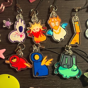 Puede incluir: Colección de coloridos colgantes acrílicos con varios diseños de dibujos animados, incluyendo un sol, un ángel y formas abstractas. Los colgantes están sujetos a cordones negros y rodeados de adornos en forma de estrella y flor.