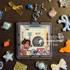 Könnte beinhalten: Ein quadratischer Acryl-Schlüsselanhänger mit einer Figur in einem weißen Raum, einem Portal-Gerät, einem Begleiter-Würfel und Roboter-Geschütztürmen. Das Design hat einen sternförmigen Metallverschluss und ist von kleinen, bunten Anhängern umgeben.
