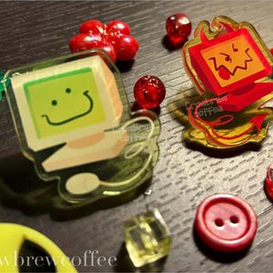 Pode incluir: Uma coleção de encantos e enfeites de acrílico coloridos. Inclui um encanto de computador verde com um rosto sorridente, um encanto de computador diabo vermelho, contas vermelhas, um botão vermelho e outros elementos decorativos. O texto "Crowbrew coffee" é visível.