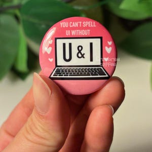 May include: A pink button with the text "YOU CAN'T SPELL UI WITHOUT U & I" in white. The button features a laptop graphic with the letters "U & I" on the screen. Small white hearts are scattered around the edges of the button.