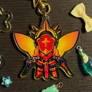 May include: A colorful acrylic charm featuring a stylized knight in red and gold armor, with a cross emblem on the helmet. The charm has a gold-colored clasp and is surrounded by other decorative charms. The text "Crowne Cafe" is visible.