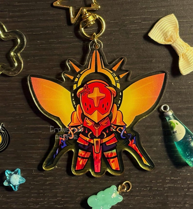 May include: A colorful acrylic charm featuring a stylized knight in red and gold armor, with a cross emblem on the helmet. The charm has a gold-colored clasp and is surrounded by other decorative charms. The text "Crowne Cafe" is visible.