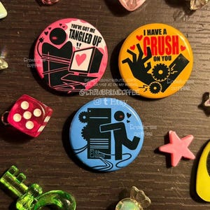 Könnte beinhalten: Drei runde Ansteckbuttons mit grafischen Illustrationen. Ein rosa Button zeigt eine Figur mit Computer und den Text "YOU'VE GOT ME TANGLED UP". Ein gelber Button zeigt eine Hand mit Zahnrädern und den Text "I HAVE A CRUSH ON YOU". Ein blauer Button zeigt eine Figur, die einen Computertower umarmt.