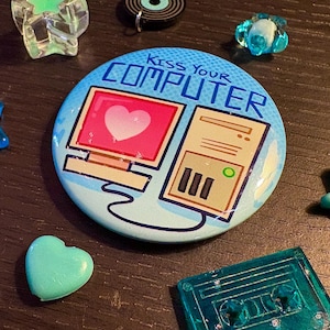 May include: A round button with the text "Kiss Your Computer" and a computer graphic with a heart on the screen. Surrounding the button are various teal-colored, star-shaped, and heart-shaped decorative items.