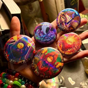 Könnte beinhalten: Fünf holografische runde Ansteckbuttons mit farbenfrohen Illustrationen und Text. Die Buttons zeigen verschiedene Charakterdesigns mit Sätzen wie "HELL IS FULL" und "BLOOD IS FUEL". Die Buttons haben eine glänzende, reflektierende Oberfläche.