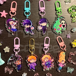 May include: A selection of anime-style acrylic keyrings, each with a unique character design. The keyrings feature colourful illustrations and are attached to metal clasps. The keyrings are in various colours, including pink, blue, green, and gold.