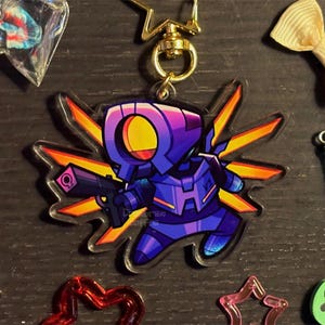 Peut inclure: Un porte-clés en acrylique transparent représentant un personnage de robot stylisé dans des tons de bleu, de violet et de jaune, tenant une arme à feu. Le porte-clés est attaché à un fermoir doré. Autour du porte-clés se trouvent divers petits objets décoratifs.