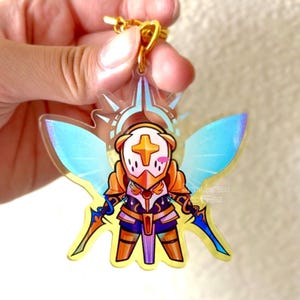 May include: A clear acrylic keychain featuring a cartoon character in gold armor, holding two blue swords. The character has a cross on its helmet and blue wings. The keychain has a gold-colored clasp.
