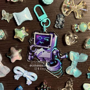 May include: An acrylic keyring featuring a purple and white robot character with a screen face displaying a smug expression, attached to a light blue plastic lobster clasp, surrounded by various small, translucent, light blue decorative charms.