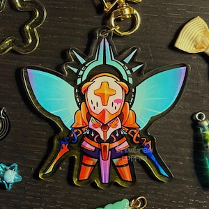 May include: A clear acrylic keychain featuring a cartoon character in armor with a cross on its helmet. The character has blue and orange accents, with iridescent wings and two swords. A gold-colored clasp is attached.