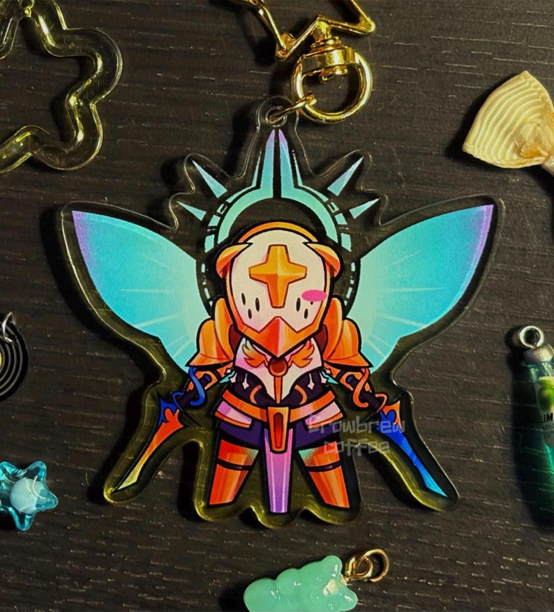 May include: A clear acrylic keychain featuring a cartoon character in armor with a cross on its helmet. The character has blue and orange accents, with iridescent wings and two swords. A gold-colored clasp is attached.