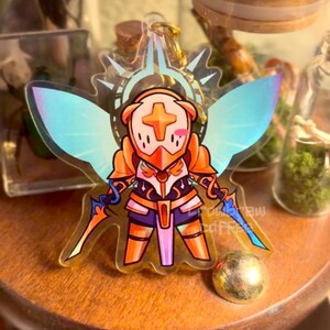 May include: A clear acrylic charm featuring a cartoon character in orange armor with a cross emblem, wings, and two swords. The charm has a gold-colored keychain attachment. The character has a blush on its cheeks.