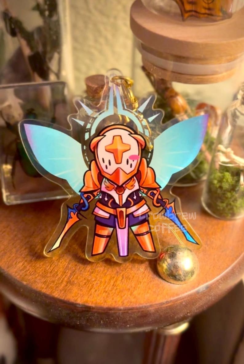 May include: A clear acrylic charm featuring a cartoon character in orange armor with a cross emblem, wings, and two swords. The charm has a gold-colored keychain attachment. The character has a blush on its cheeks.