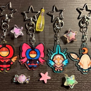 May include: Four keyrings with star-shaped clasps and character charms. The charms feature colourful designs of figures with wings, bows, and helmets. Additional star-shaped and candy-shaped charms and accessories are scattered around the keyrings.