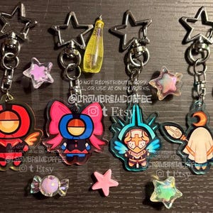 May include: Four acrylic keychains with star-shaped clasps featuring stylised character illustrations in red, blue, and white. The designs include a red winged figure, a blue figure with large bows, a knight-like figure, and a cloaked figure with a staff.