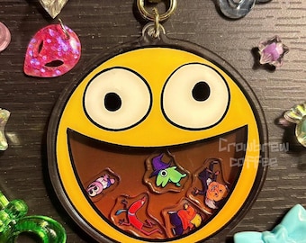 Könnte beinhalten: Ein gelber Smiley-Schlüsselanhänger mit einem braunen Mund, der mit bunten Cartoon-Figuren gefüllt ist. Der Schlüsselanhänger hat einen goldfarbenen sternförmigen Verschluss und ist von verschiedenen dekorativen Anhängern umgeben.
