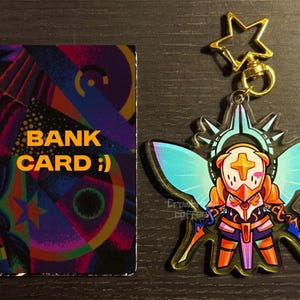 May include: A colorful bank card with the text "BANK CARD :)" next to a charm featuring a character with wings, a gold star, and a gold clasp. The charm has orange, blue, and purple accents.
